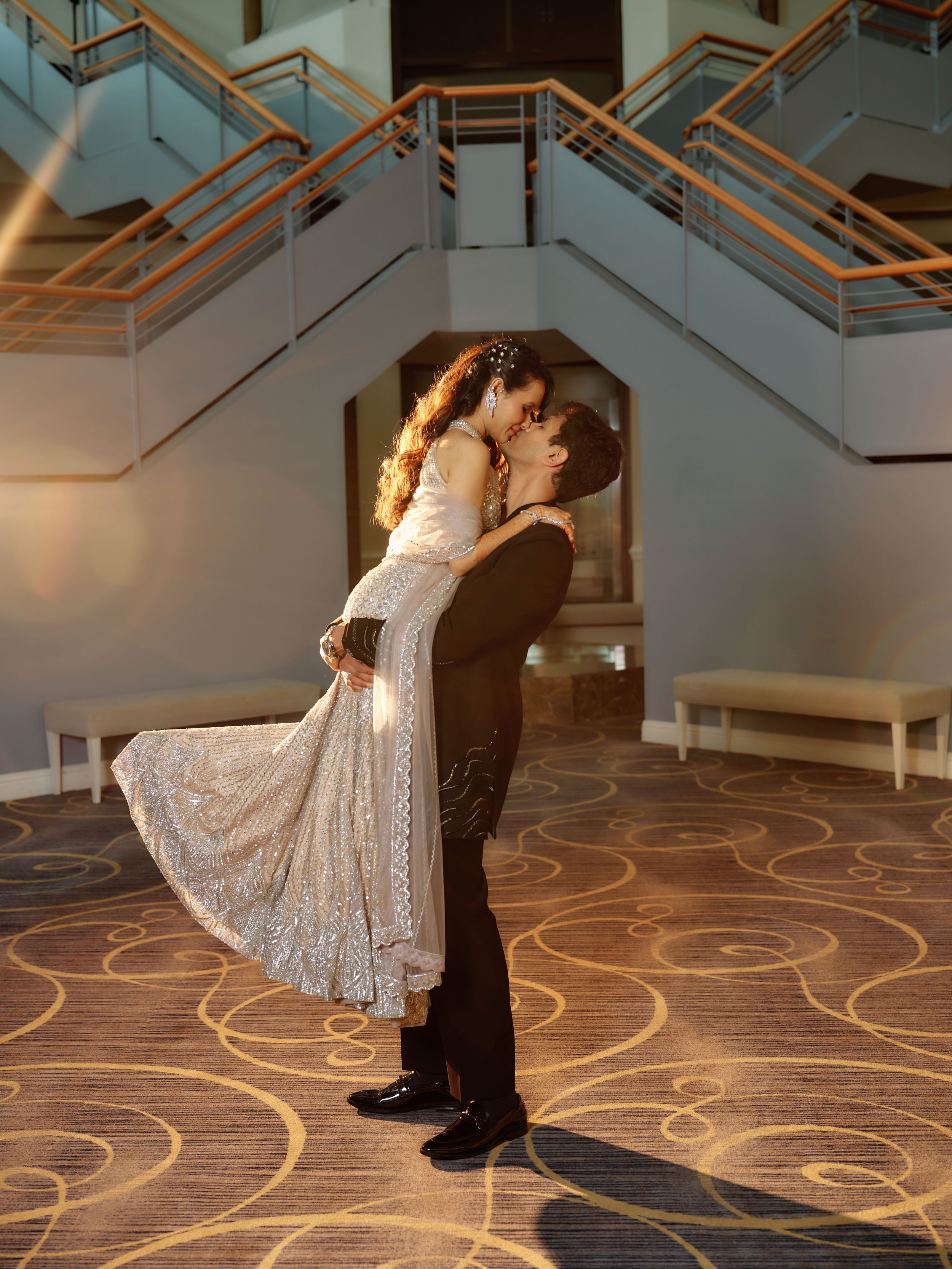 Bride in white lace dress lifted by groom in tuxedo sharing romantic kiss in elegant hotel lobby.