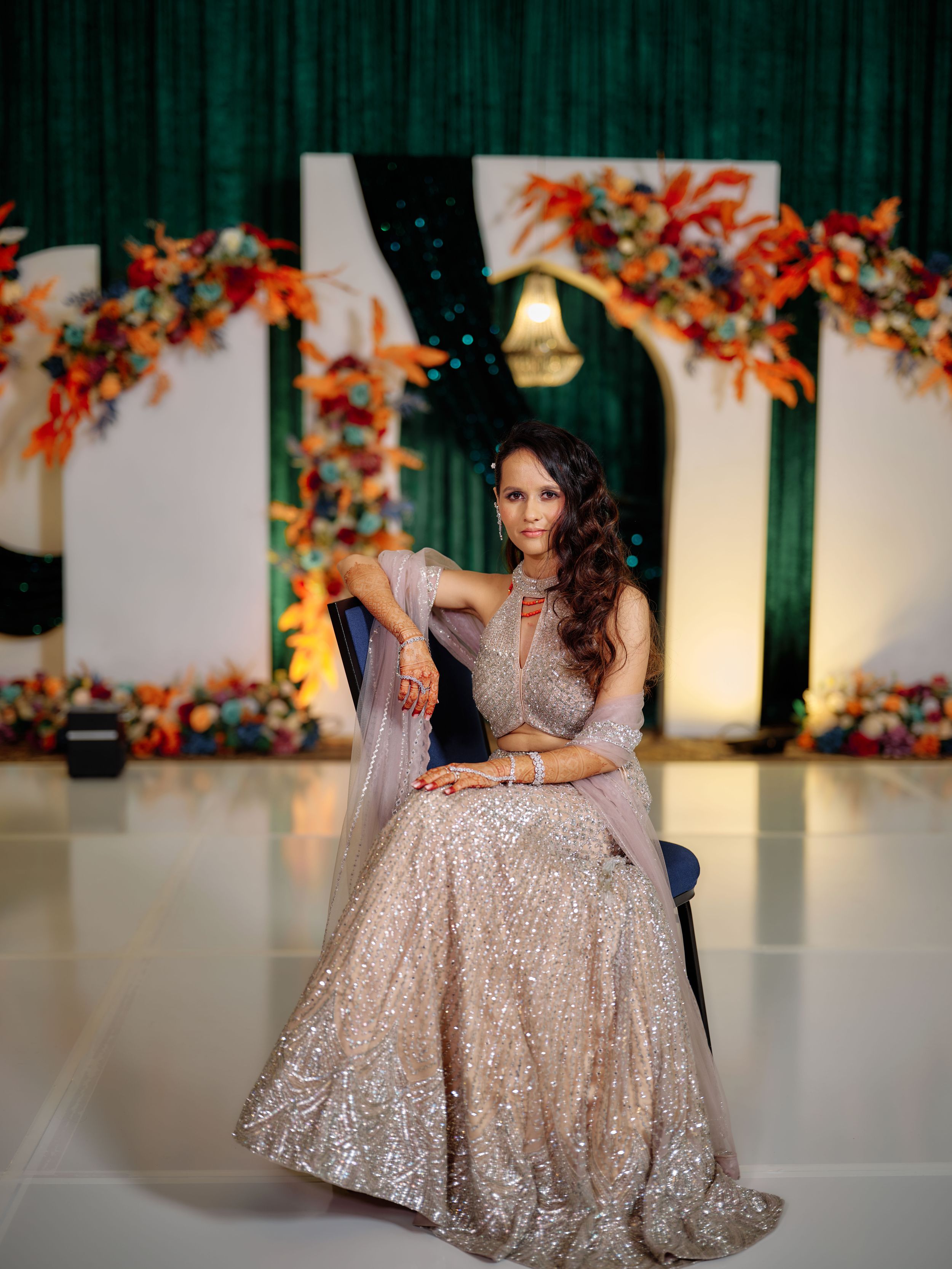 Woman in sparkly champagne lehenga sits elegantly before green backdrop with orange floral decor.