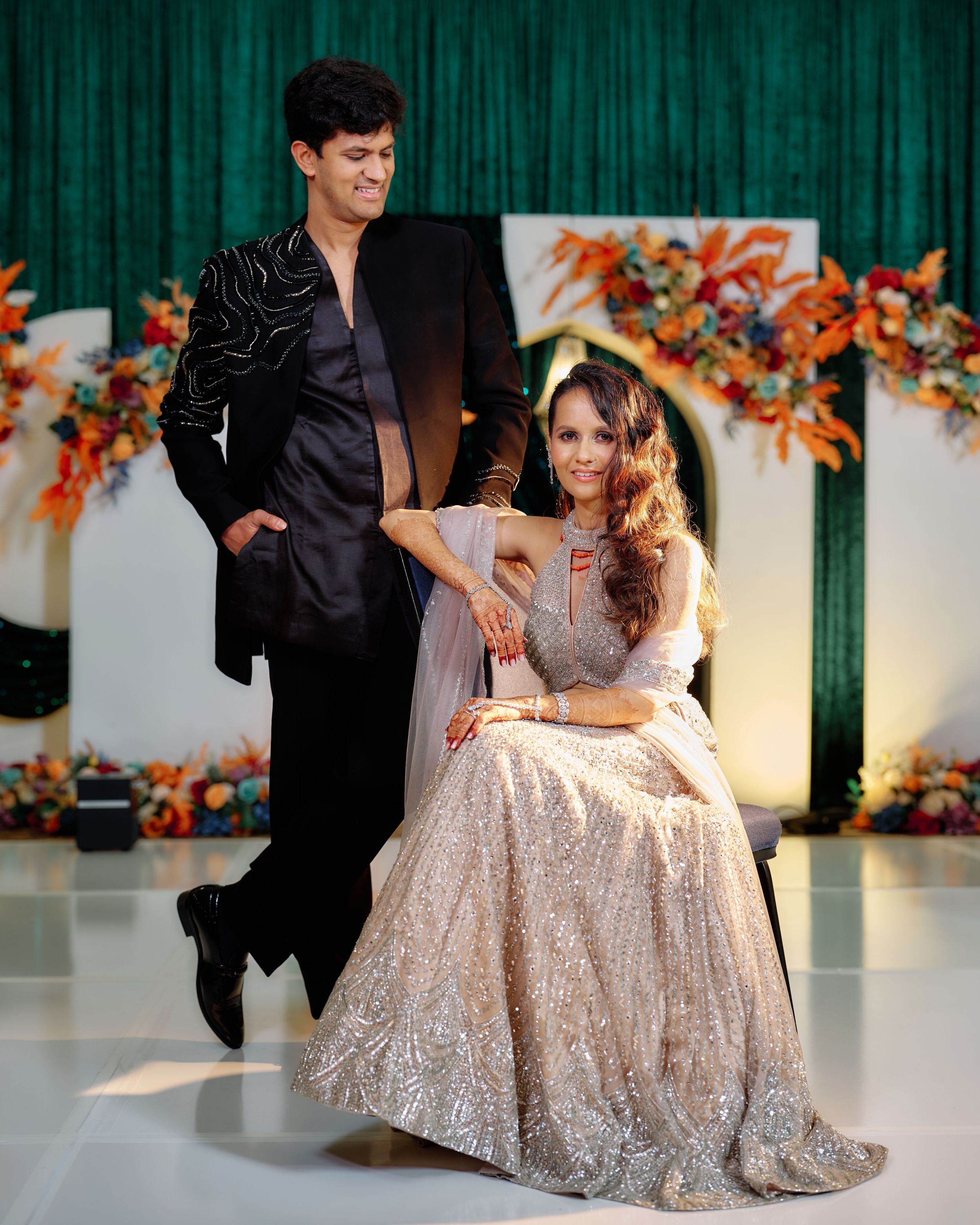 Couple poses elegantly on white dance floor, woman in gold lehenga seated, man in black suit standing behind her.