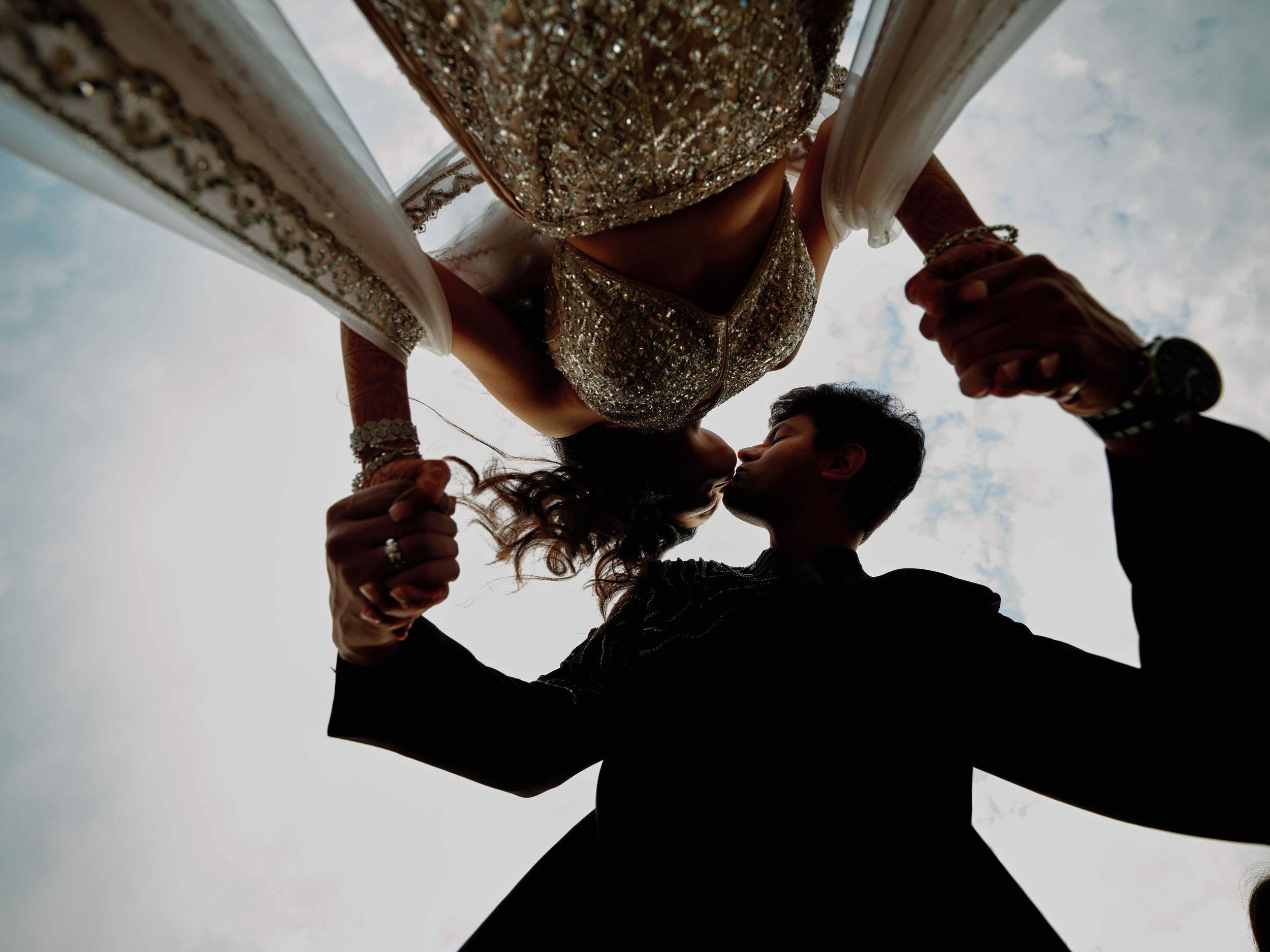 Upward angle shot of couple sharing a kiss under ornate wedding canopy, groom in black suit, bride in embroidered gown.