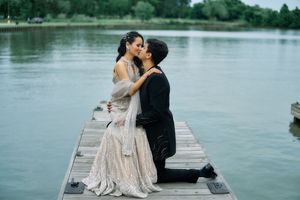Couple sharing a romantic kiss on a wooden dock by a serene lake, bride in a shimmering gown, groom kneeling in a suit.