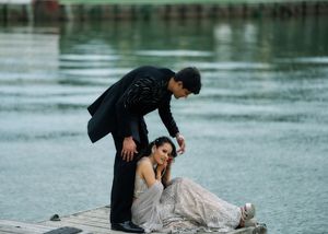 A couple in elegant formal wear poses on a dock, man in black suit standing over woman in silver gown seated gracefully.