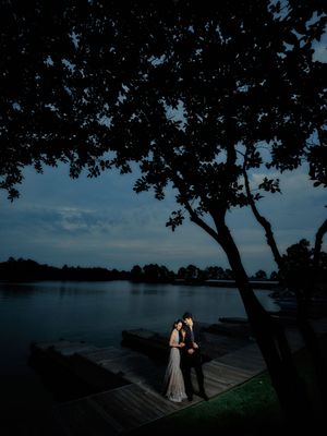 Couple embraces on a lakeside dock at dusk, framed by silhouetted trees under a dramatic blue twilight sky.
