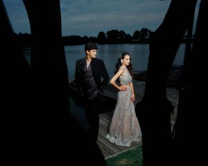 Elegant couple in formal attire posing on a waterfront dock at dusk, man in black suit, woman in embroidered gown.