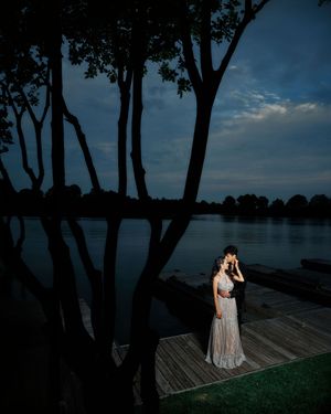 Romantic couple embracing on a lakeside dock at dusk, surrounded by tall trees reflecting on calm water.