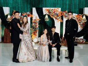Family posing elegantly at a festive South Asian celebration with green sequin backdrop and colorful floral decor.