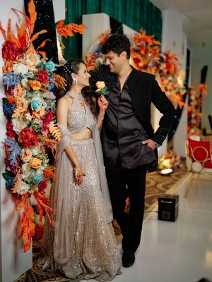 Couple posing at Indian wedding, woman in silver lehenga, man in black suit, colorful floral backdrop.