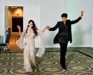 Couple dancing joyfully in ballroom, woman in silver lehenga and man in black kurta holding hands mid-step.