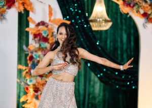 Bride in silver lehenga dancing gracefully in front of vibrant floral and emerald green backdrop.
