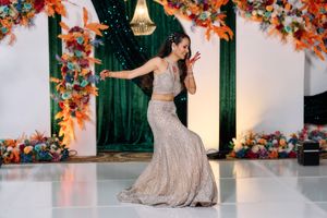 Woman performs solo dance on white floor in sparkly gold lehenga skirt before colorful floral backdrop.
