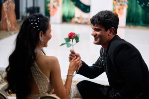 A man kneeling and offering a red rose to a woman in a gold dress at a romantic formal event.