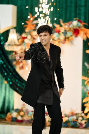 Man in black embroidered sherwani pointing at camera against green sequin and floral wedding backdrop.