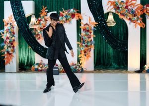 Performer in black suit dancing on white floor with colorful floral backdrop and green drape decor.
