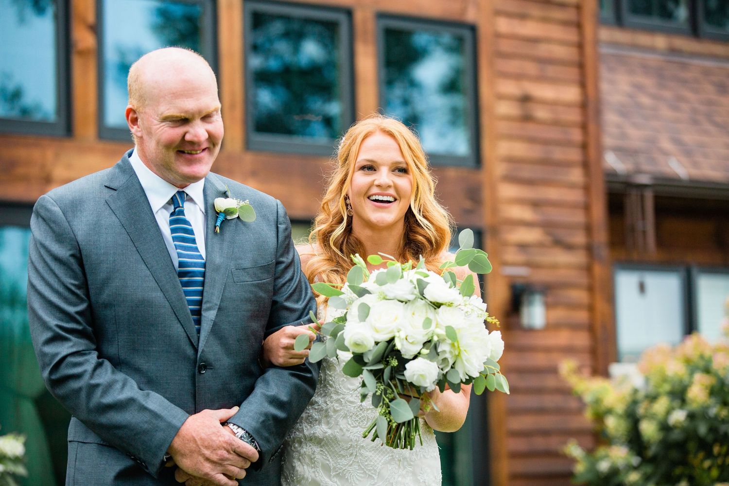 Smiling bride holding white bouquet walks with groom in gray suit outside rustic wooden wedding venue.