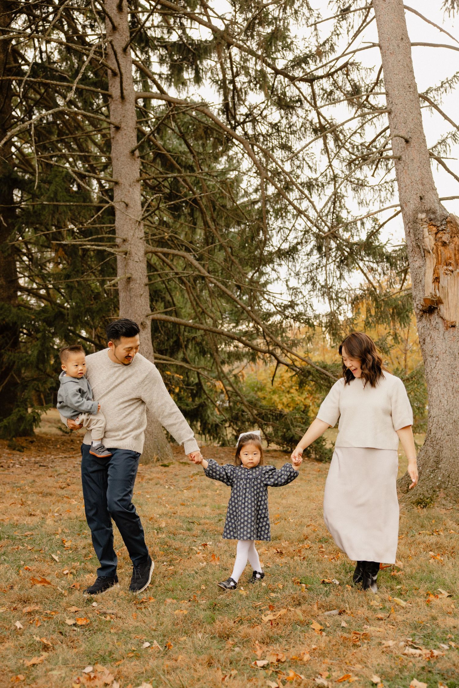 Family of four walking outdoors in autumn park, parents holding toddler girl's hands while dad carries baby boy.