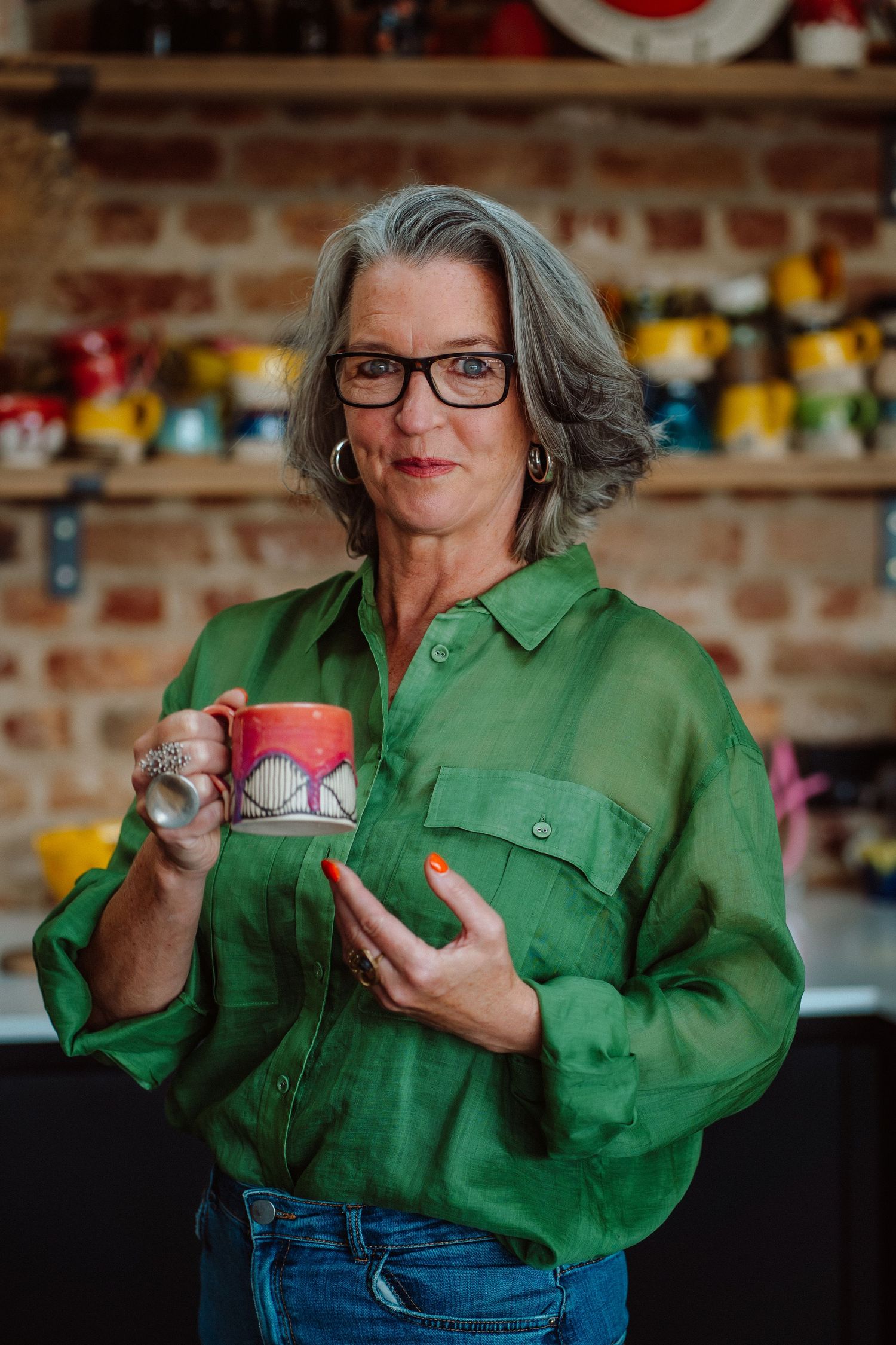 Middle-aged woman with gray hair and glasses holding a colorful mug, wearing a green shirt in a rustic kitchen.
