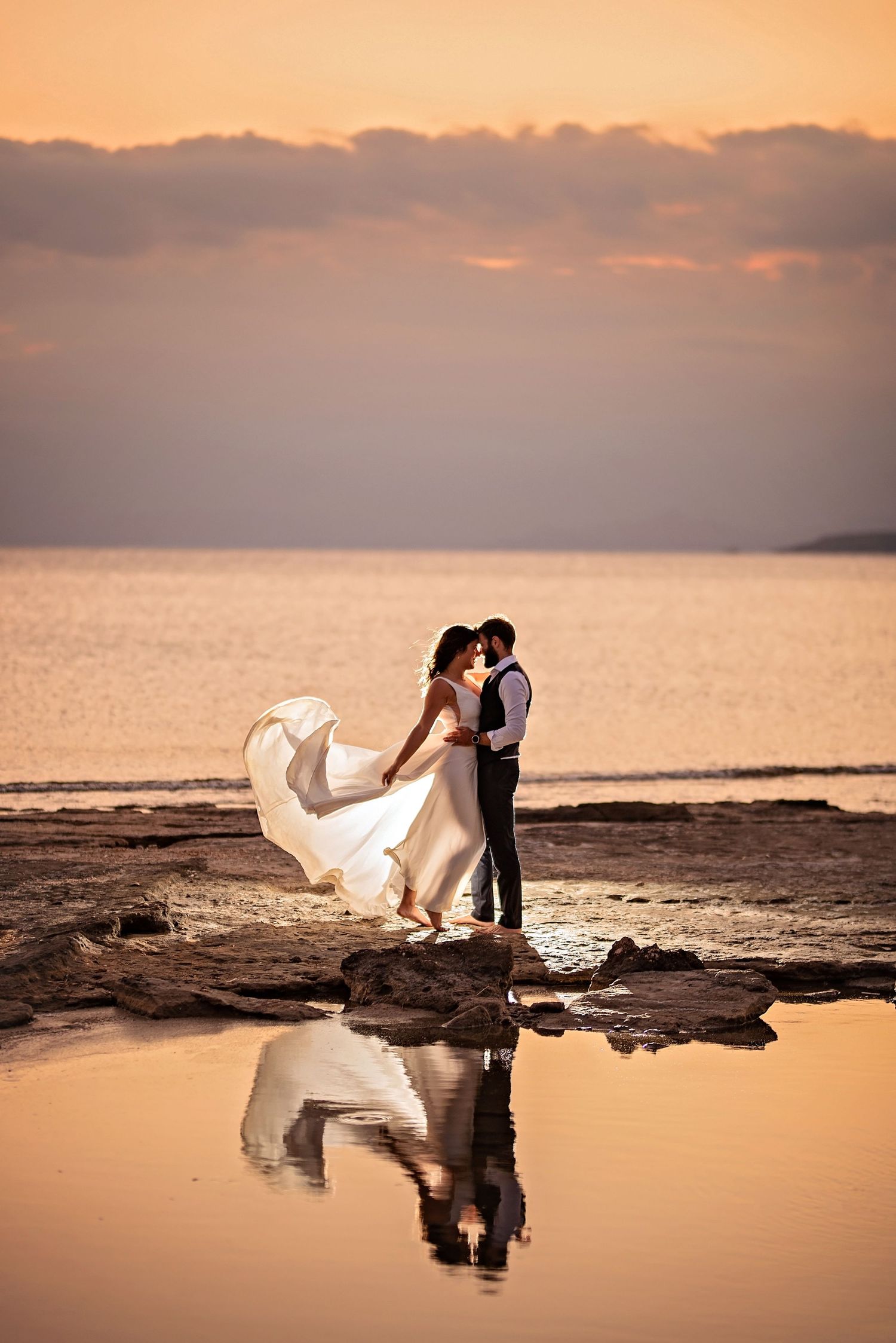 Bride and groom embrace at sunset on rocky shore, dress flowing in wind, with romantic reflection in water below.