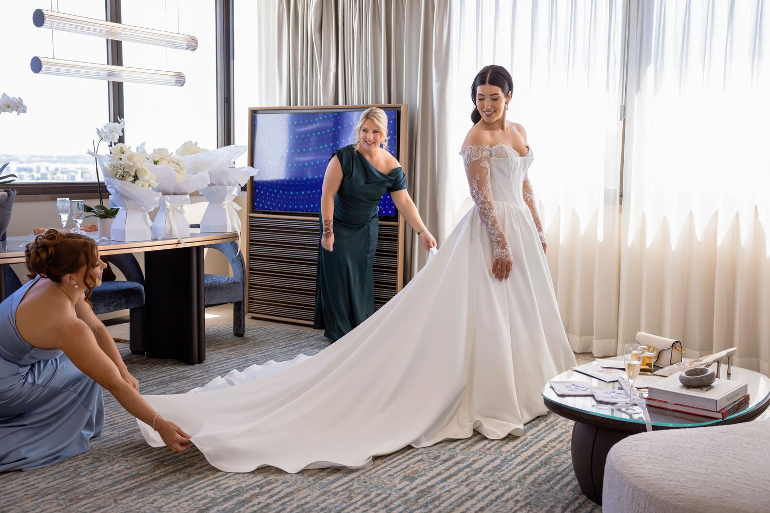 Bride in elegant white ball gown being helped by bridesmaids in blue and green dresses in a hotel room.