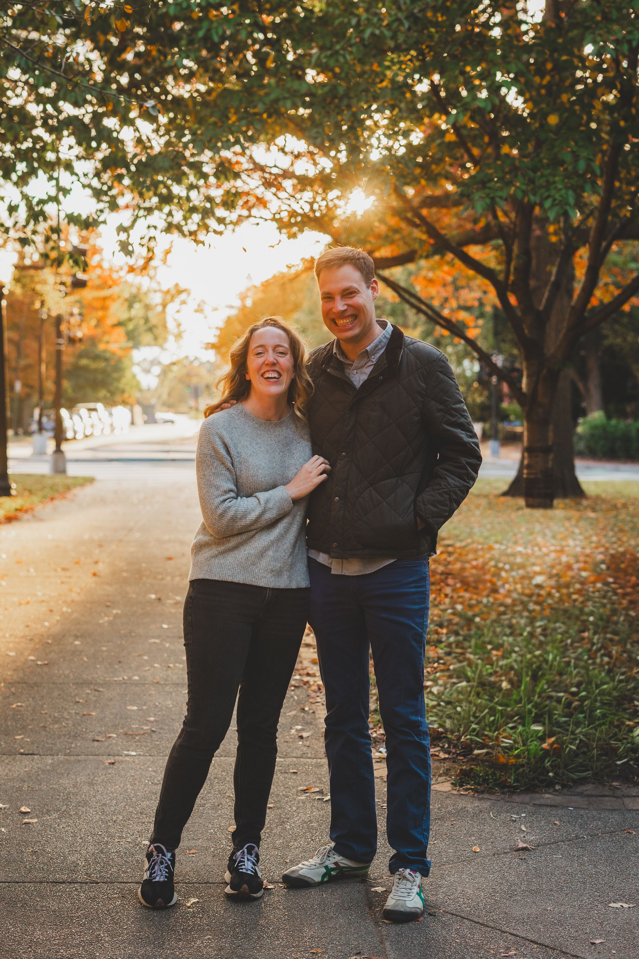 Couple smiling together on a tree-lined path during golden autumn sunset, woman in gray sweater and man in dark jacket.