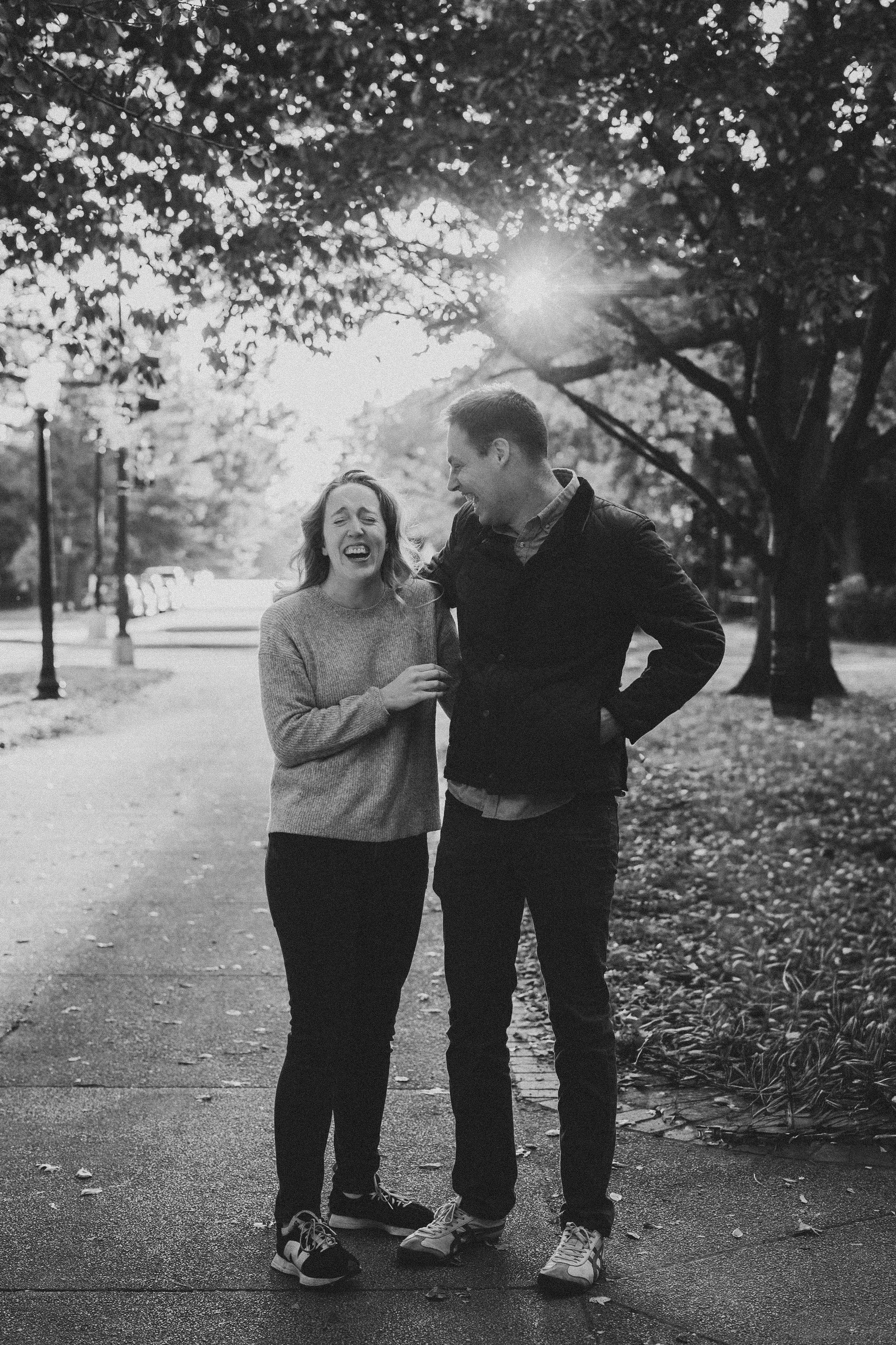 Black and white photo of a laughing couple standing together on a tree-lined path in a park.