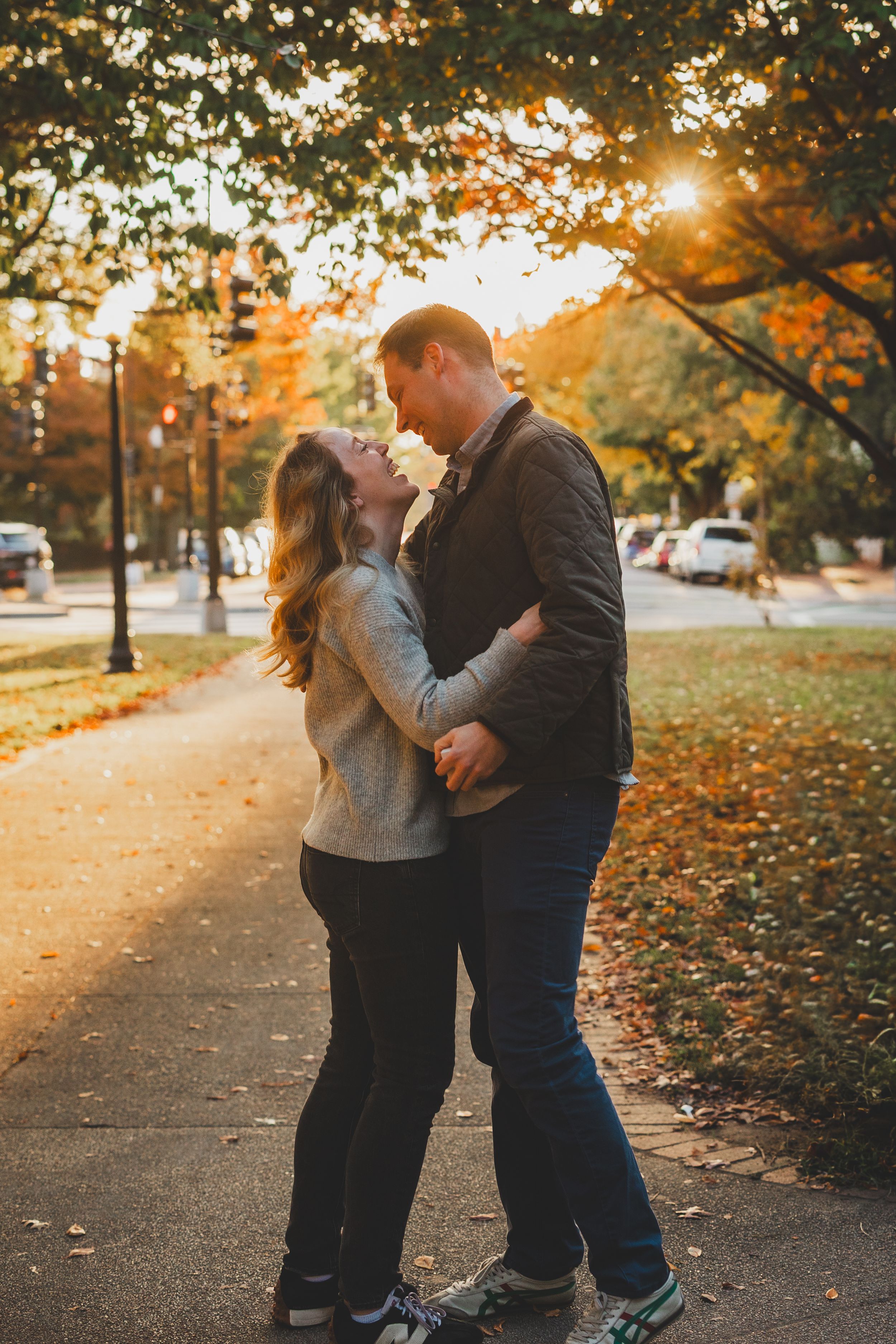Couple embracing and gazing at each other in a sunlit autumn park with golden foliage and a tree-lined path at Stanton Park In DC
