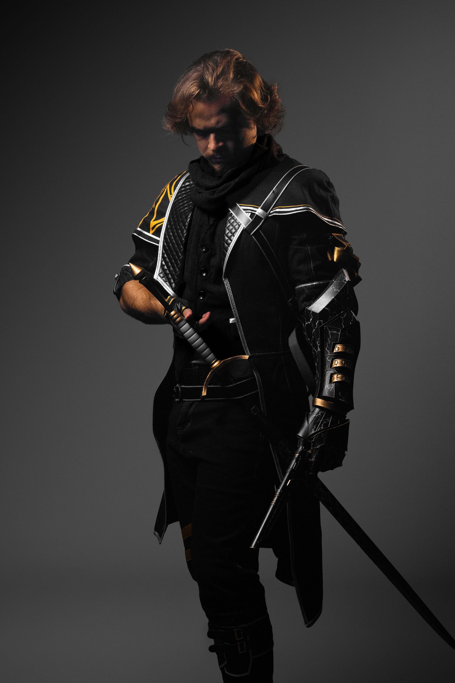 A warrior in black and gold armor holds a katana sword, wearing dark fantasy cosplay costume against gray background.