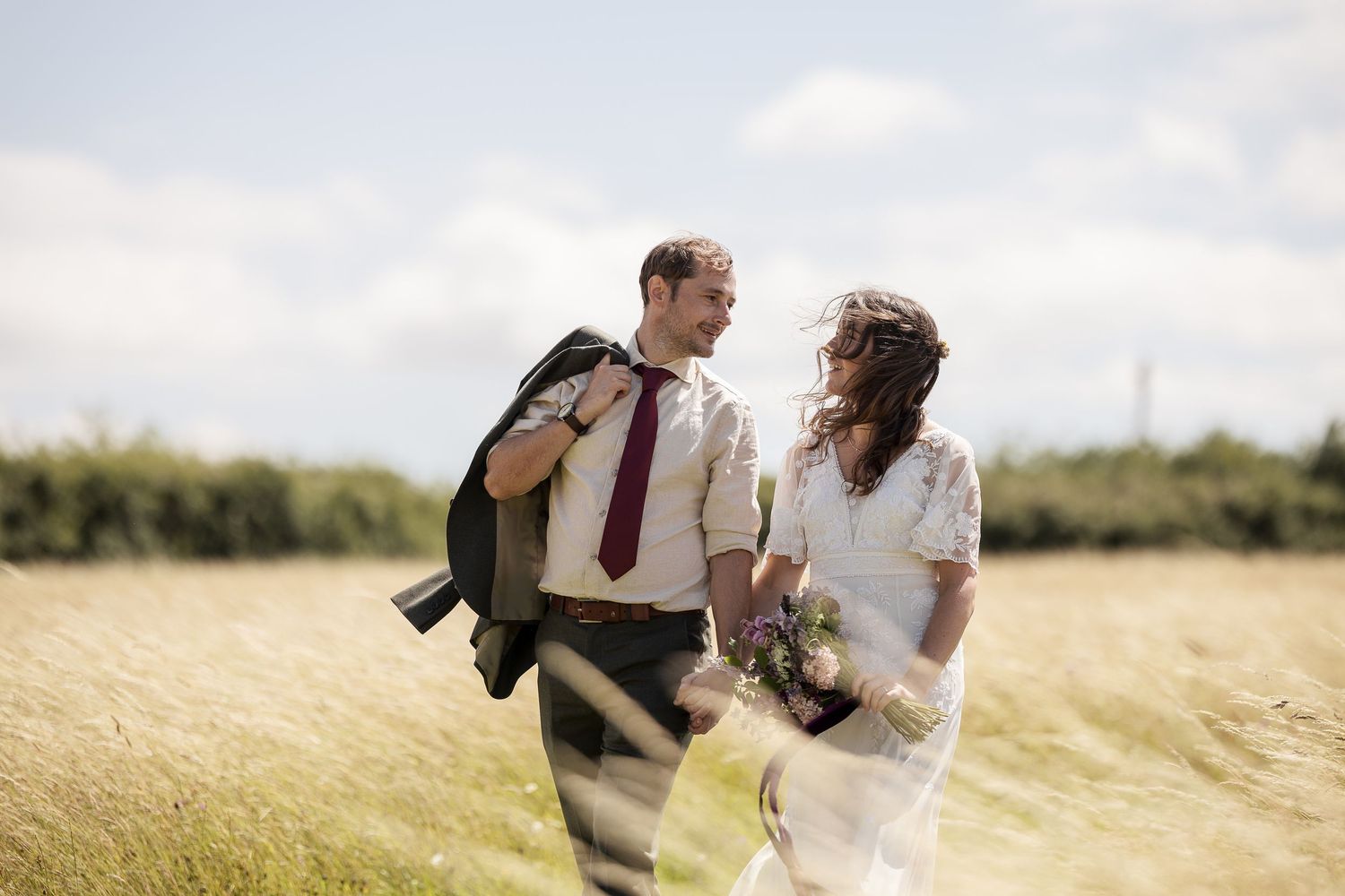 A happy couple walks through a sunny field, the bride holding wildflowers in a white dress, the groom in a white shirt and red tie.