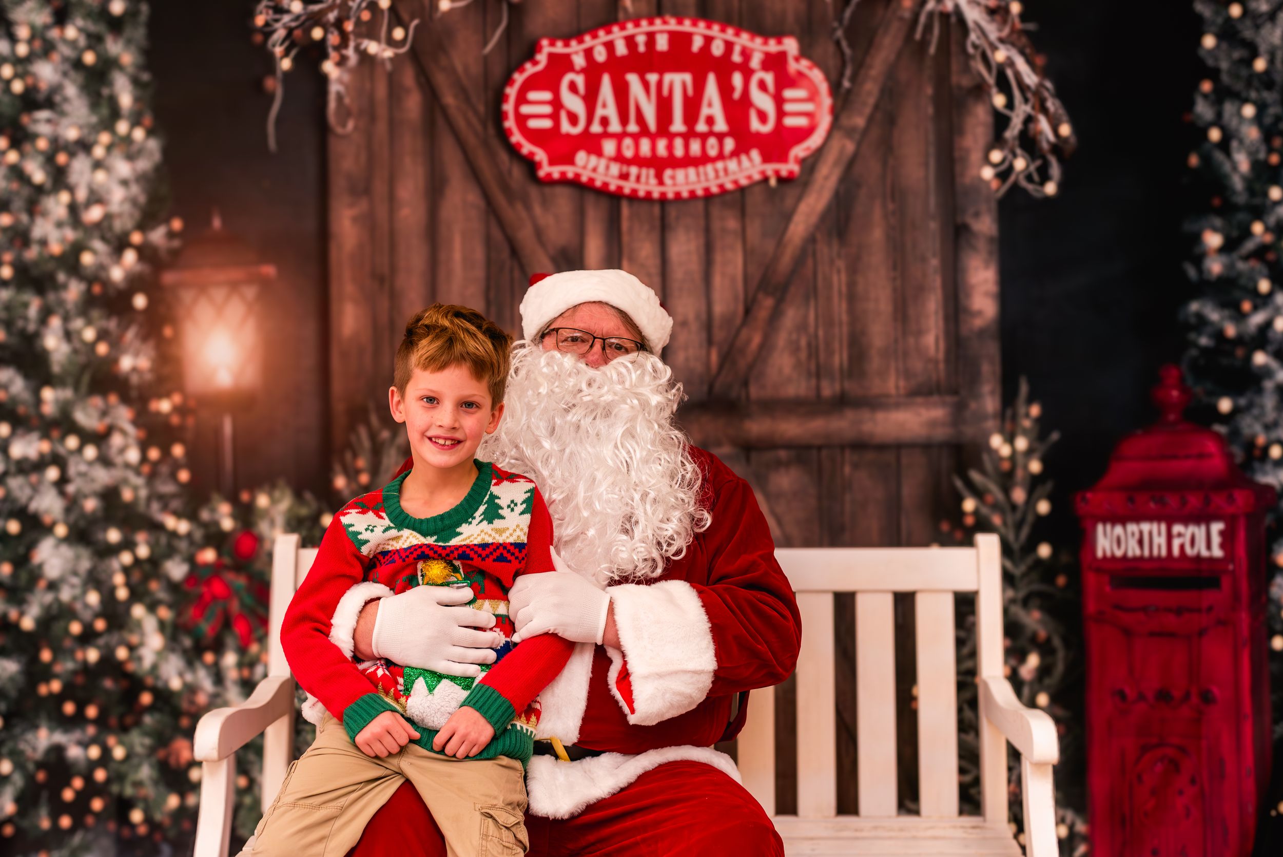 Two children sit with Santa Claus in his workshop, surrounded by festive Christmas decorations and trees.
