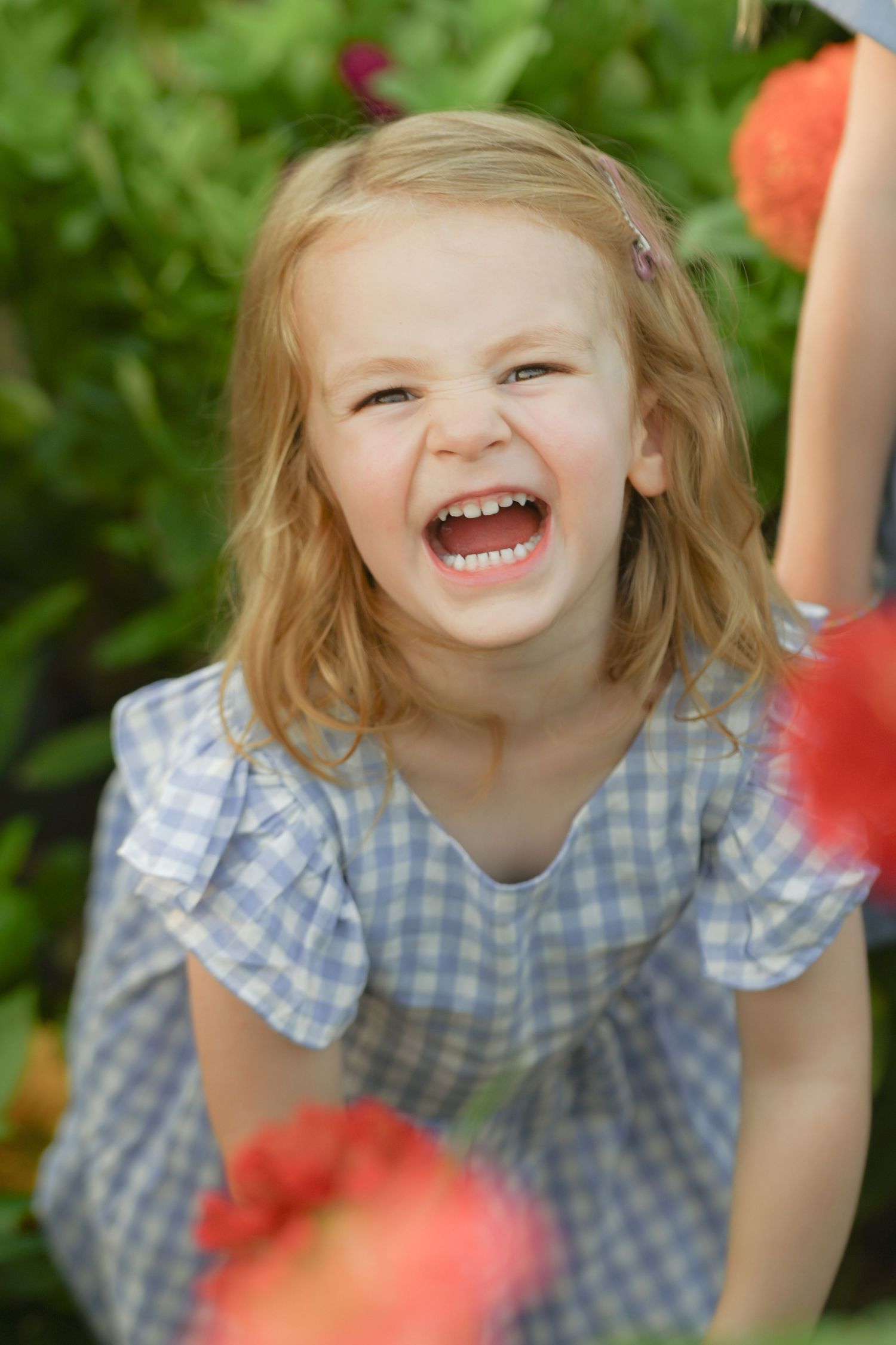 Joyful young blonde girl in blue gingham dress laughing outdoors among colorful red flowers and green foliage.