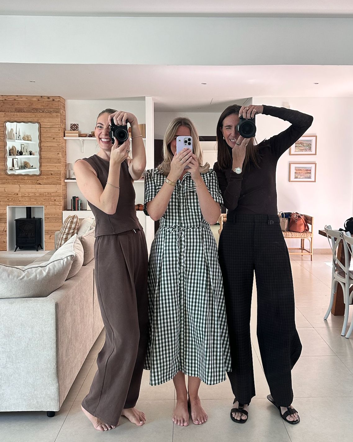 Three women pose in a mirror selfie, wearing stylish outfits in brown, gingham, and black, holding cameras in a modern living room.