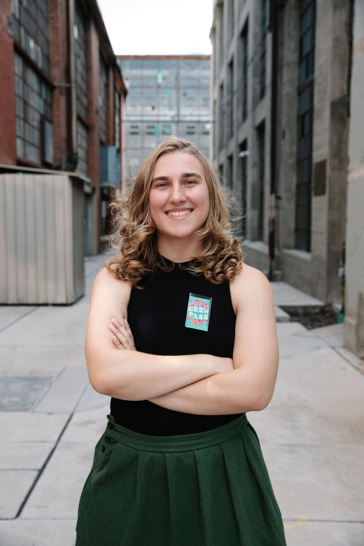 Young woman with curly blonde hair smiling in urban alley, wearing black top and green skirt with arms crossed.