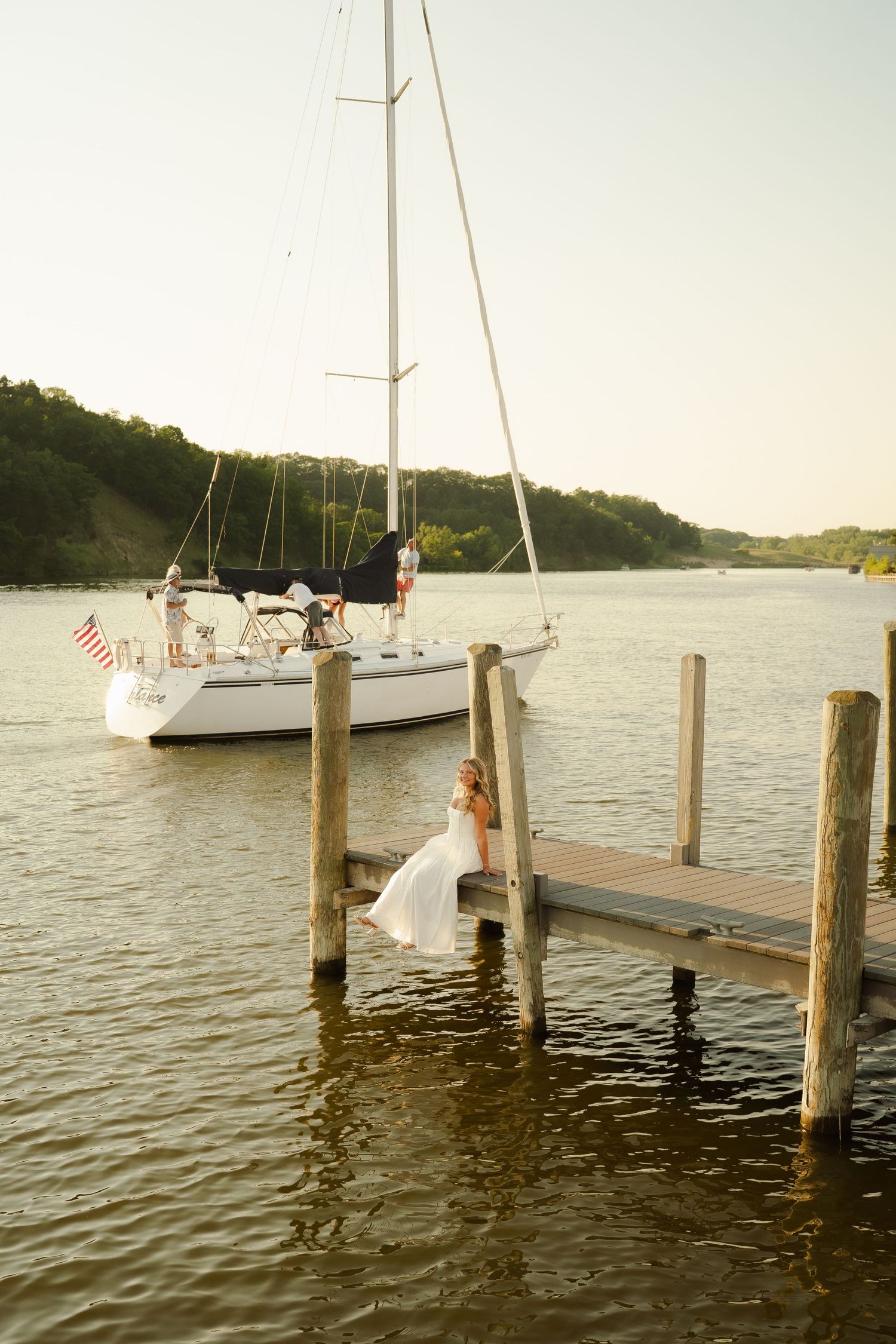 Bride in white gown poses on wooden dock beside sailboat on calm river at golden hour.