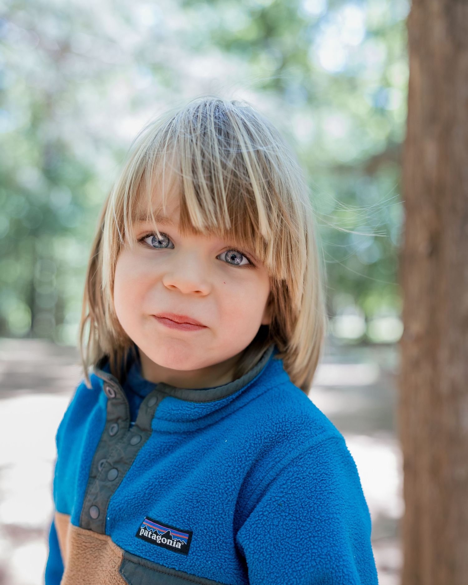 A young blonde child with blue eyes wearing a blue Patagonia fleece jacket outdoors among trees.