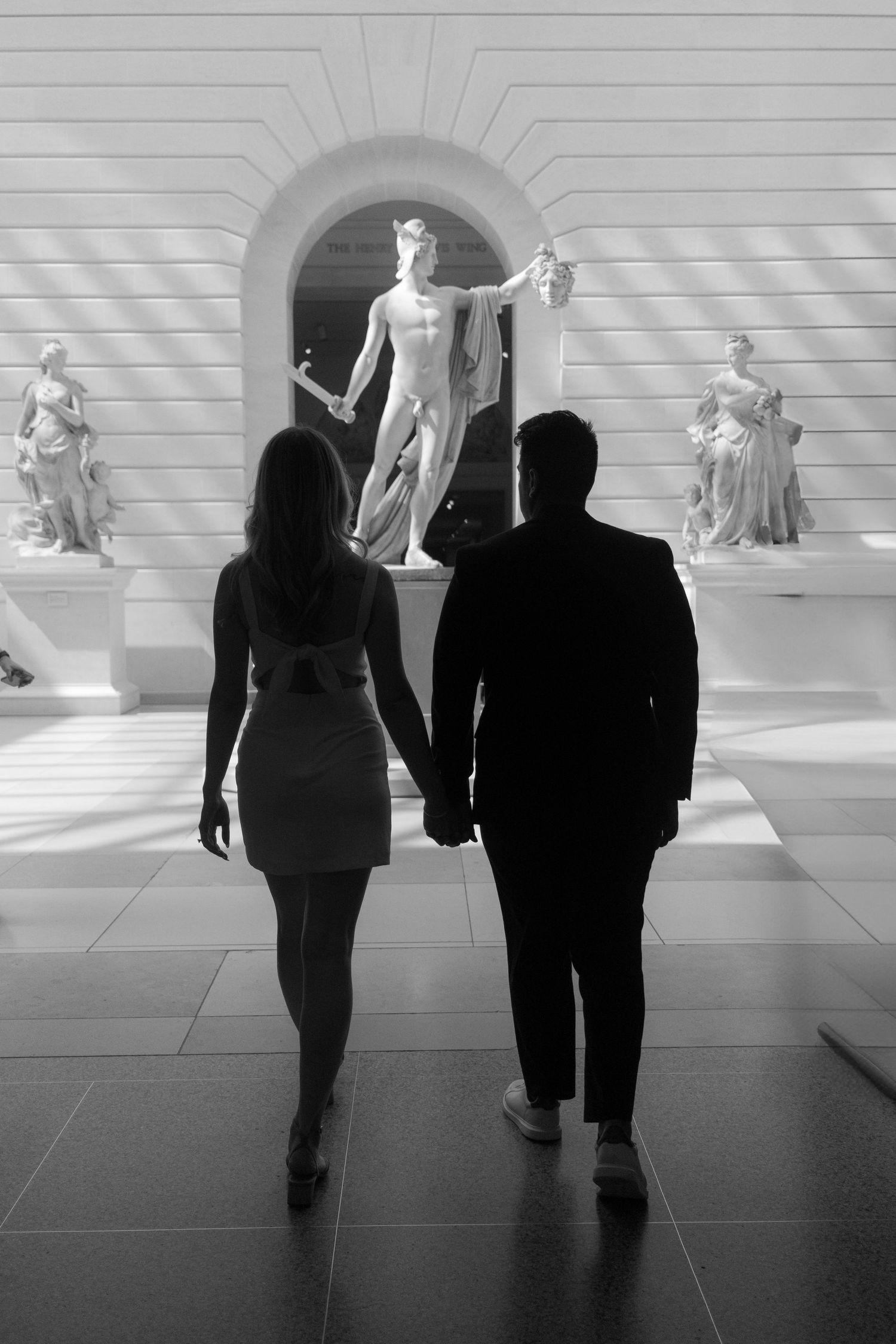 Black and white photo of a couple holding hands walking toward classical sculptures in an elegant museum gallery.