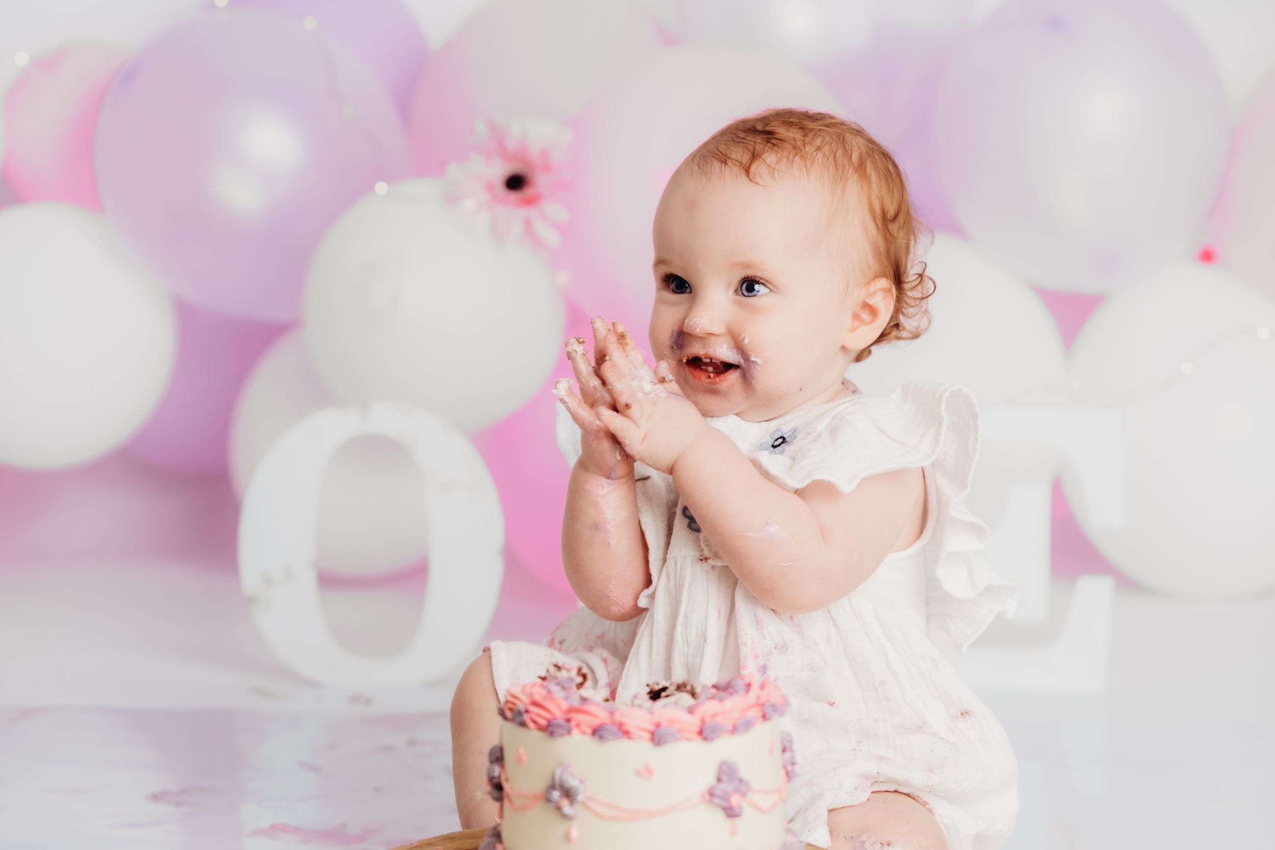 Baby in white dress claps hands next to pink birthday cake with pastel balloon backdrop.