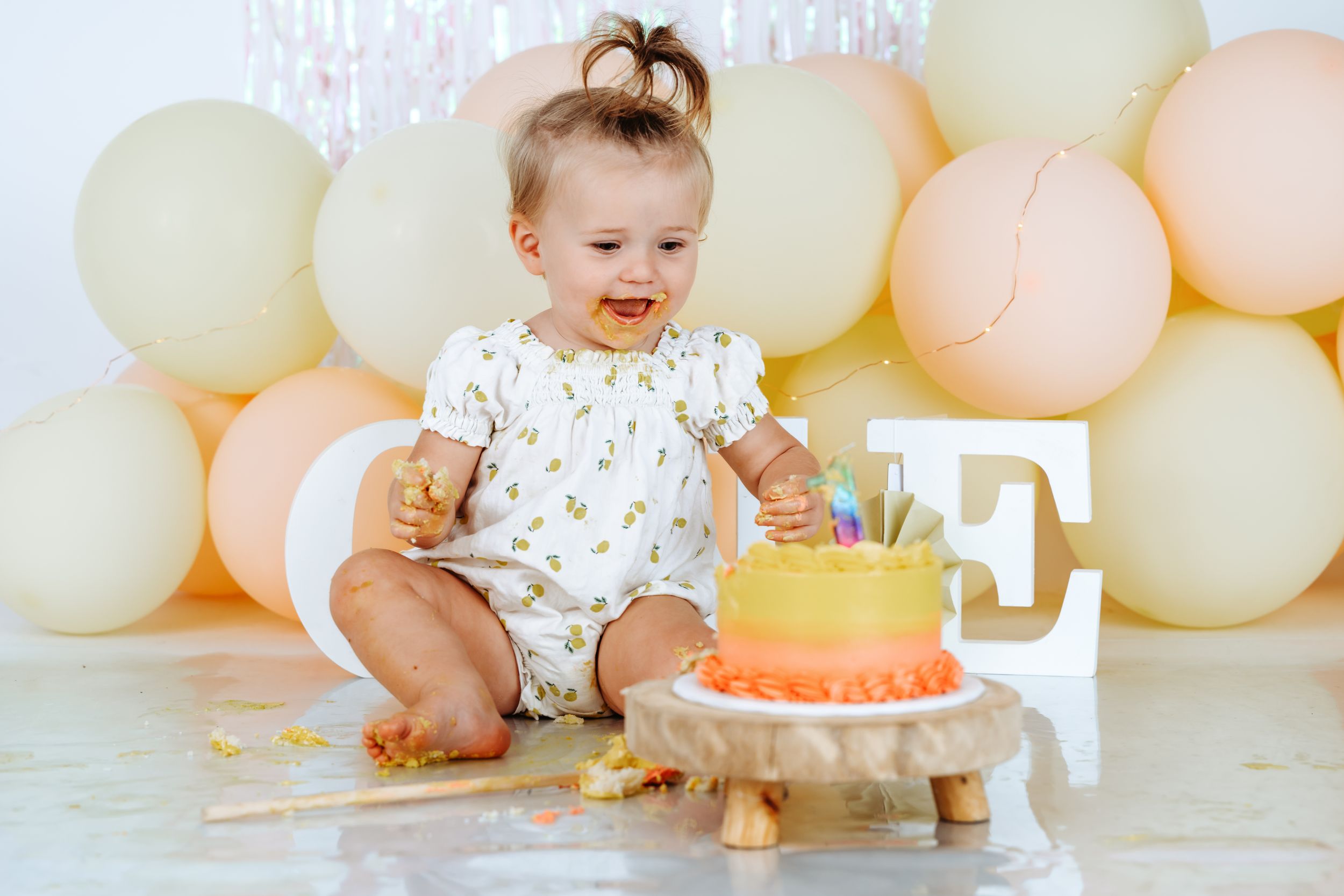 A baby in a floral outfit sits on the floor with a birthday cake and pastel balloons in the background for a first birthday celebration.