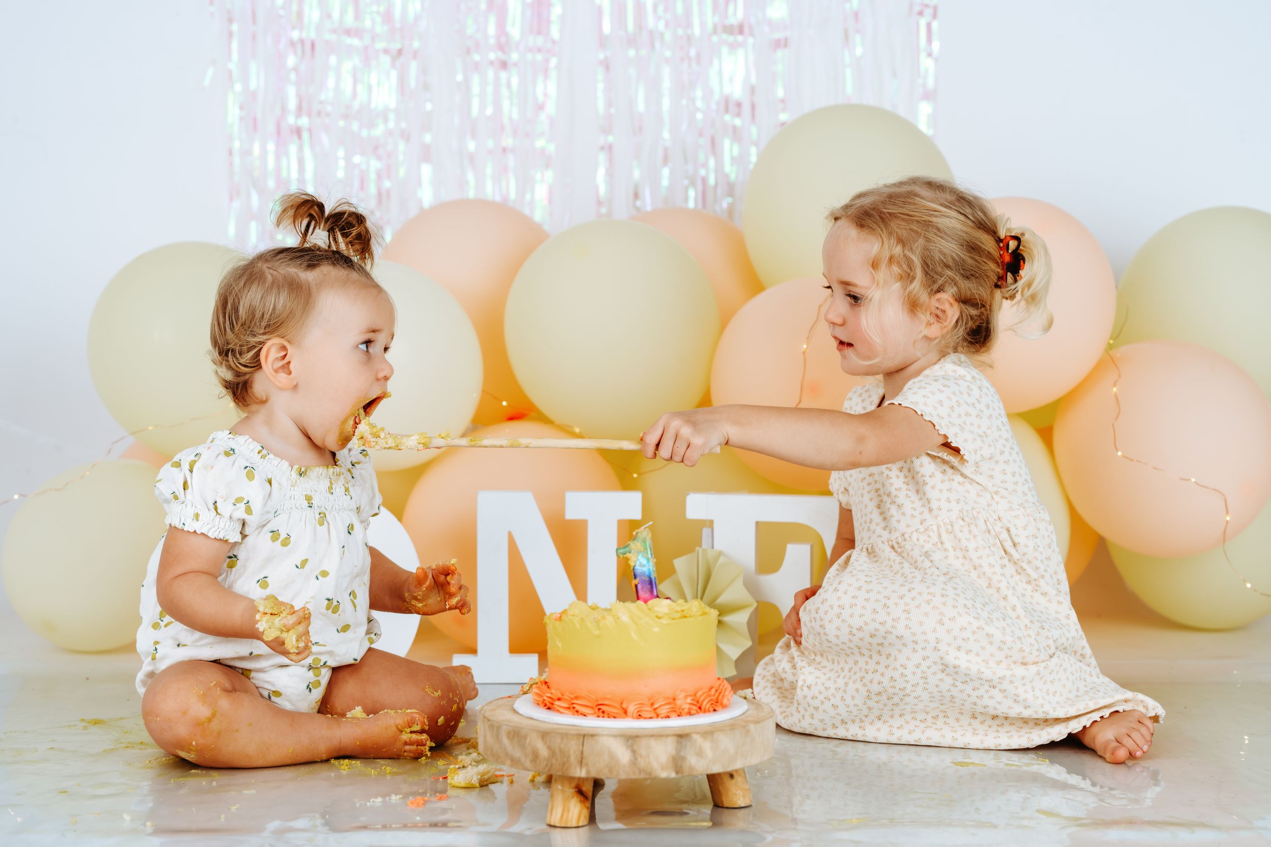 Two babies sitting on floor with birthday cake and balloons, one wearing white dress and one wearing white onesie.