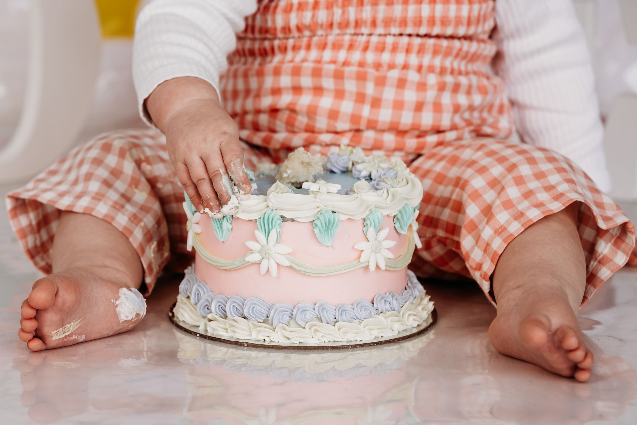 A small birthday cake decorated with daisies and pink frosting sits in front of tiny feet in a gingham dress.