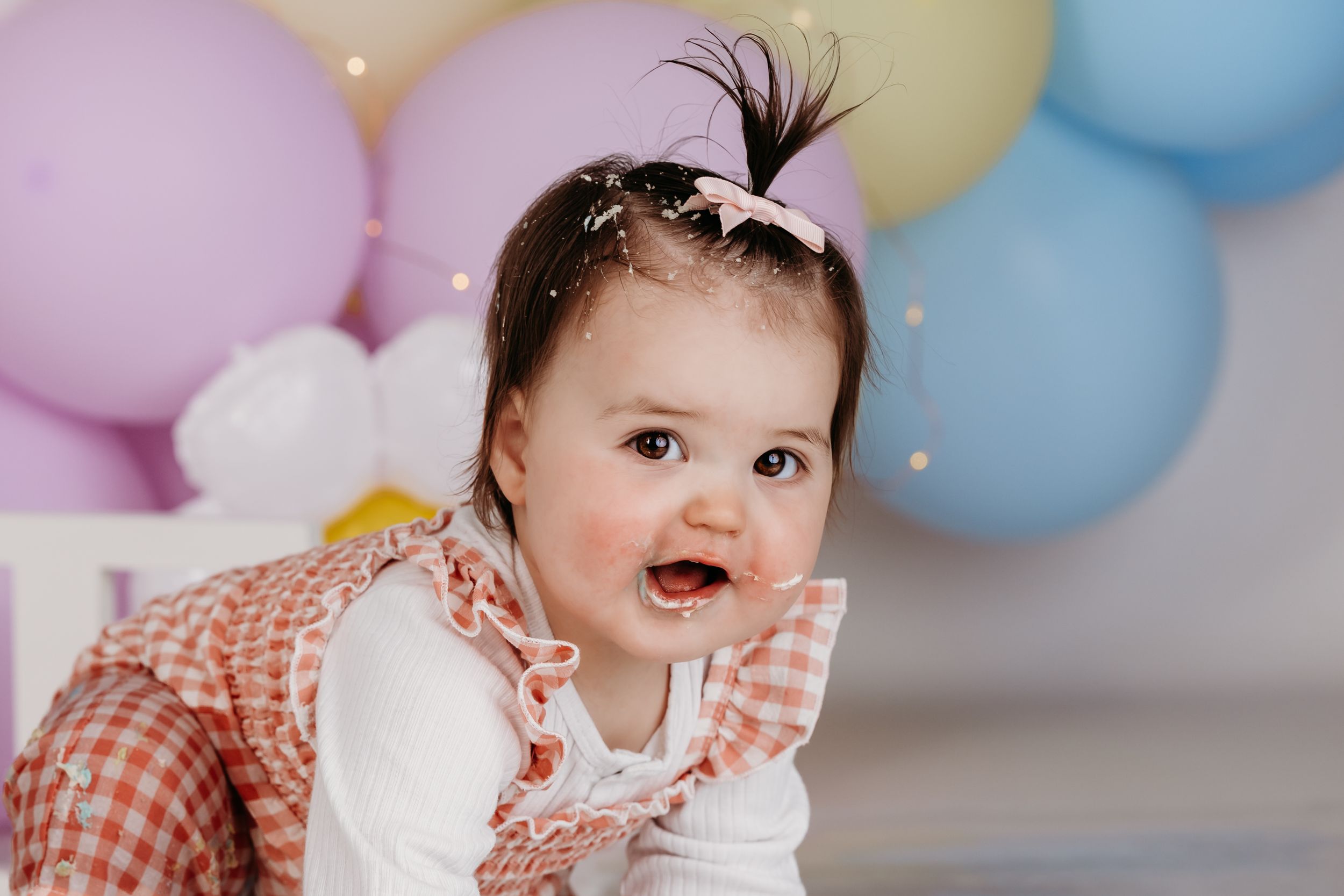 Happy baby wearing gingham dress and hair bow crawls on floor with pastel balloons in background.