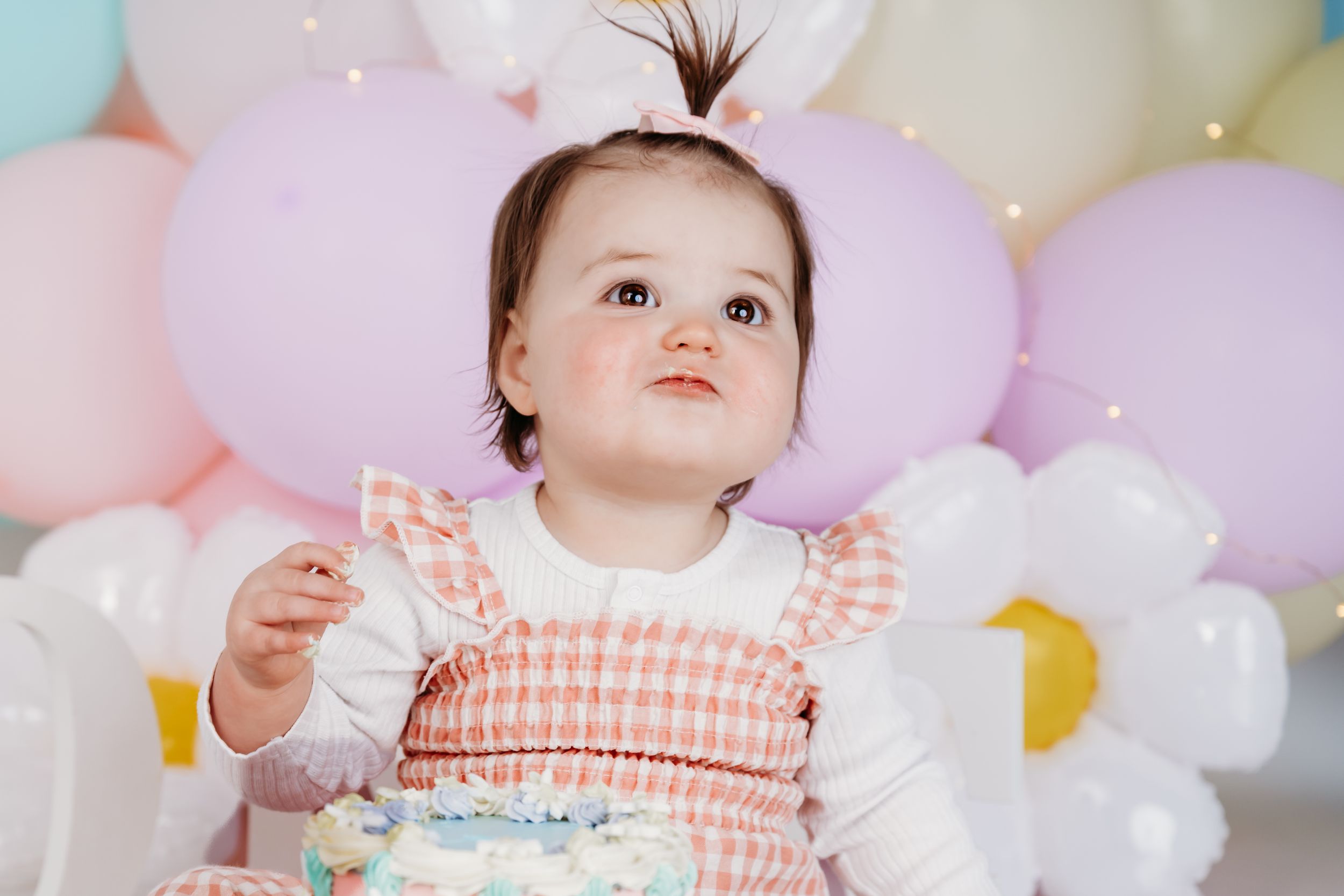 Baby wearing a gingham dress smiles upward while seated in front of pastel balloons at a birthday celebration.