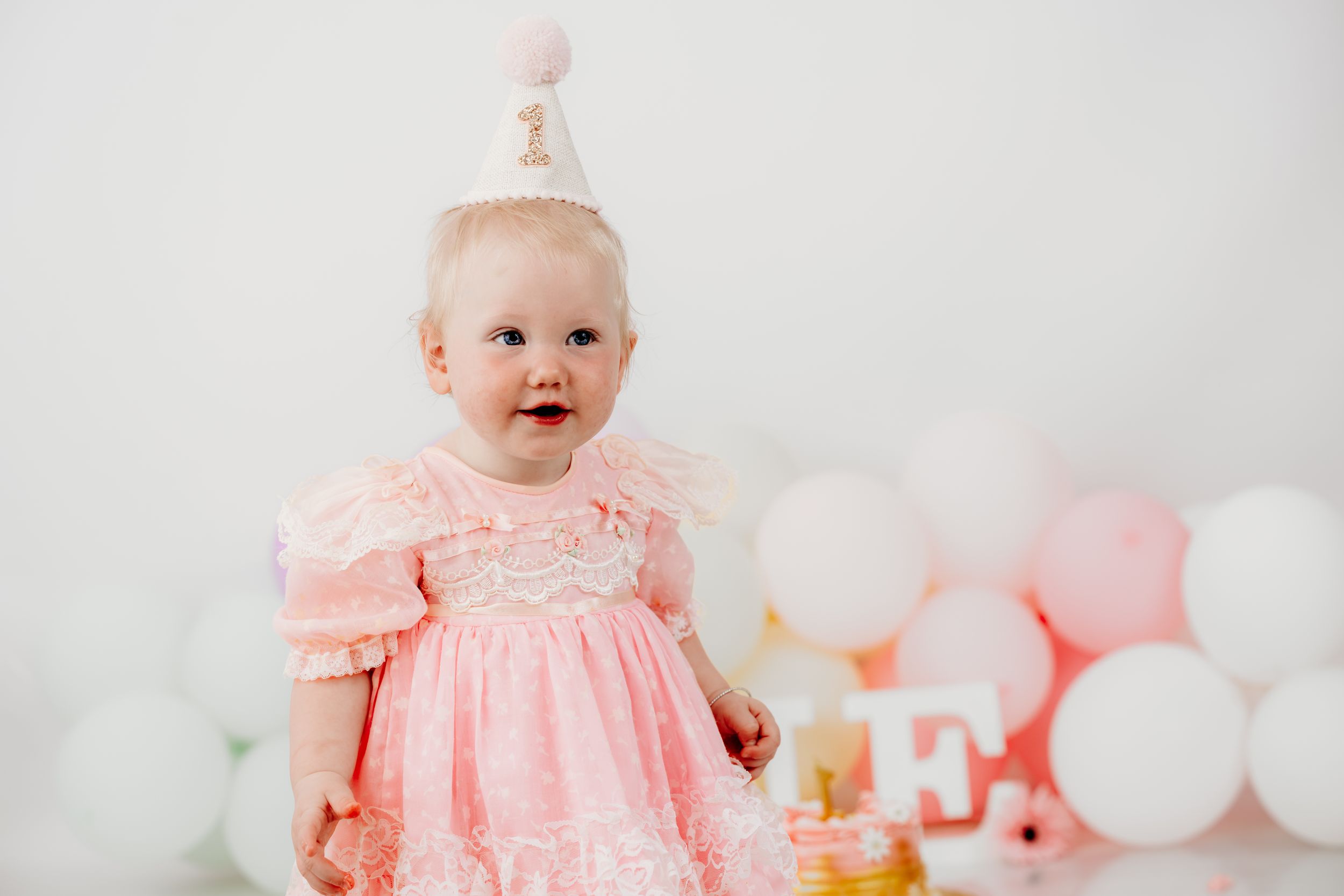 Baby wearing pink dress and party hat stands in front of white and pink balloon decorations at first birthday celebration.