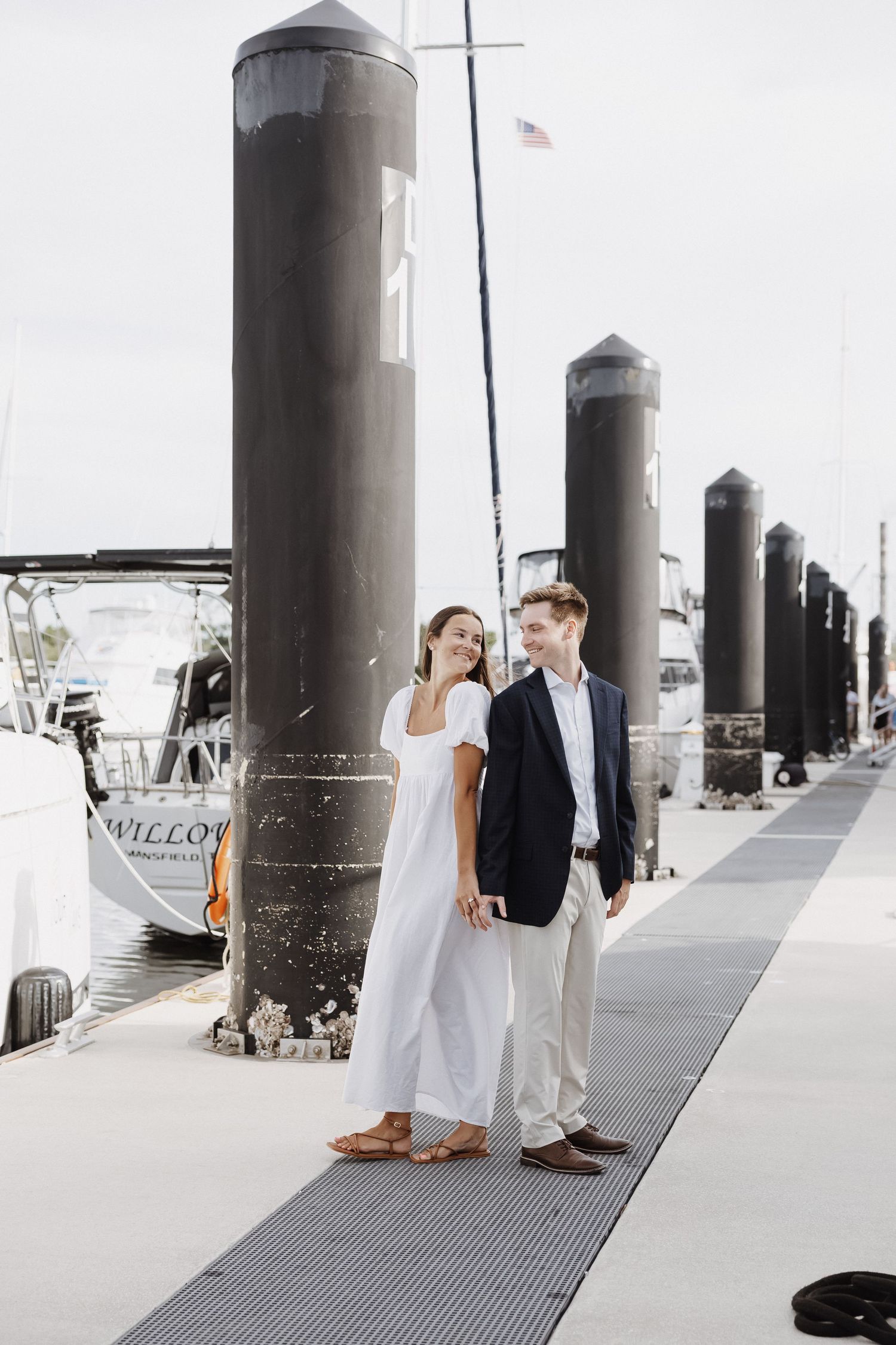 Elegant couple walking along marina dock with boats and tall black pillars in background.