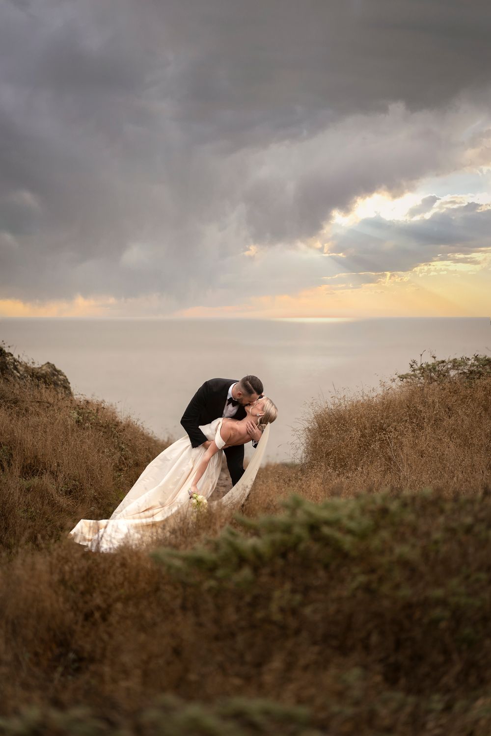 A romantic couple shares an intimate dip kiss against a dramatic stormy sky and golden grass at sunset.