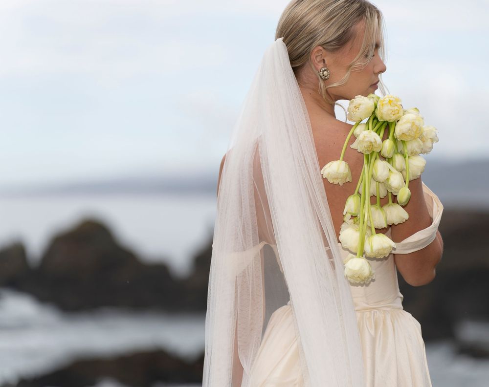 A bride in a white wedding dress holds a bouquet of yellow calla lilies against an ocean backdrop.