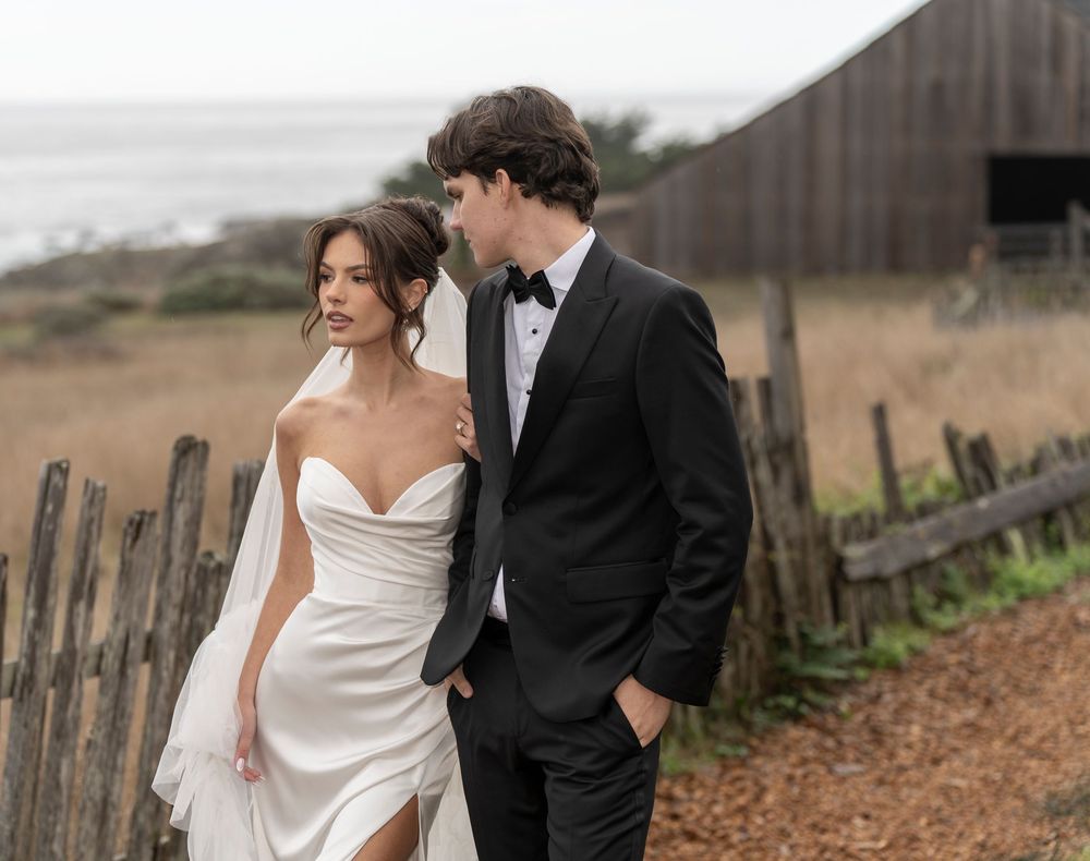 A romantic couple in wedding attire walks together along a rustic wooden fence on an autumn day.