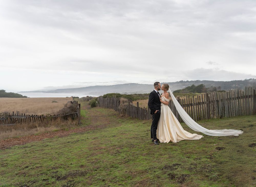 Newlyweds pose for romantic photos in a scenic coastal field with rolling hills in the background.