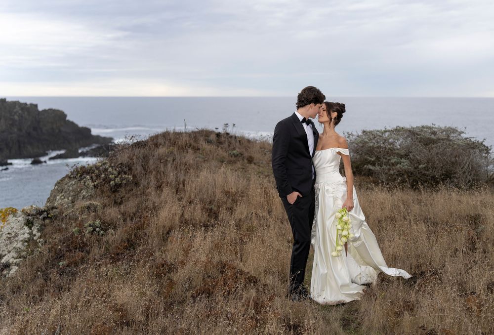 A romantic coastal wedding photoshoot featuring a couple embracing on dramatic cliffs overlooking the ocean at sunset.