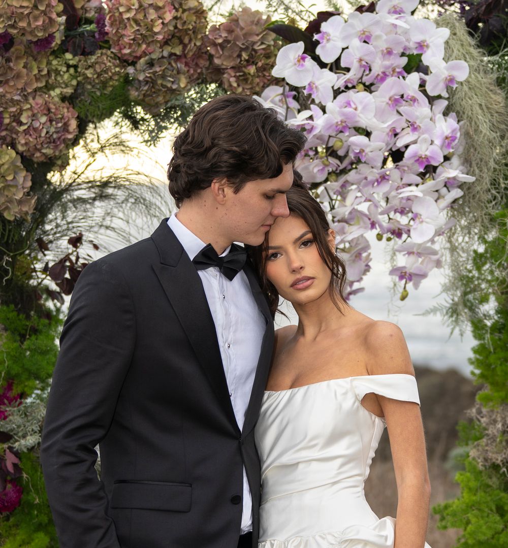 A romantic wedding couple embraces under blooming pink cherry blossoms in a garden setting.