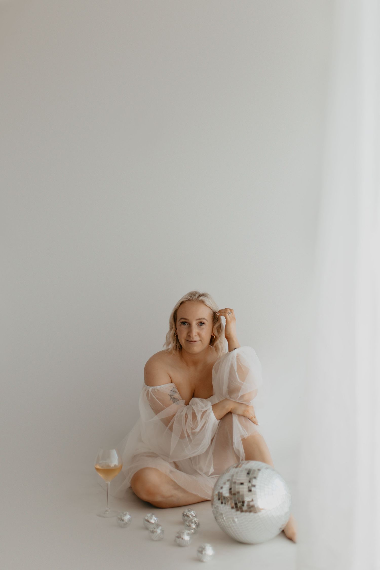 Woman in sheer dress poses with wine glass and disco ball on white background.