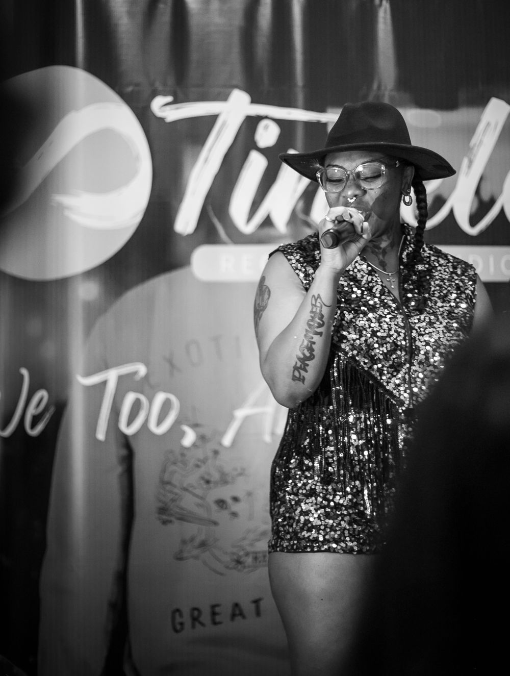 A performer in a sparkling dress and black hat sings on stage with 'Tired' signage visible in the black and white photo.